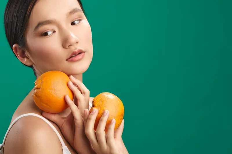 asian woman gracefully holds bright oranges against a bold backdrop in a clean studio, with soft lighting and a calm pose that radiates freshness and wellness.