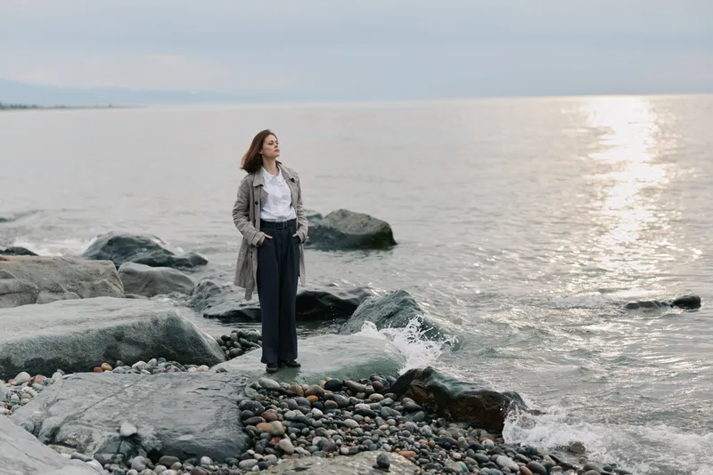 Woman stands on rocky shore near calm sea wearing long coat and trousers. Cloudy sky and reflective water create serene atmosphere. Concept of solitude, nature, and contemplation.