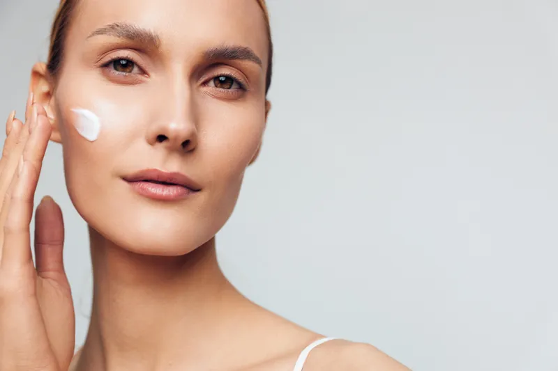 Young woman applying face cream with fingers, clear skin, natural makeup, soft lighting, neutral background, skincare routine, beauty care concept