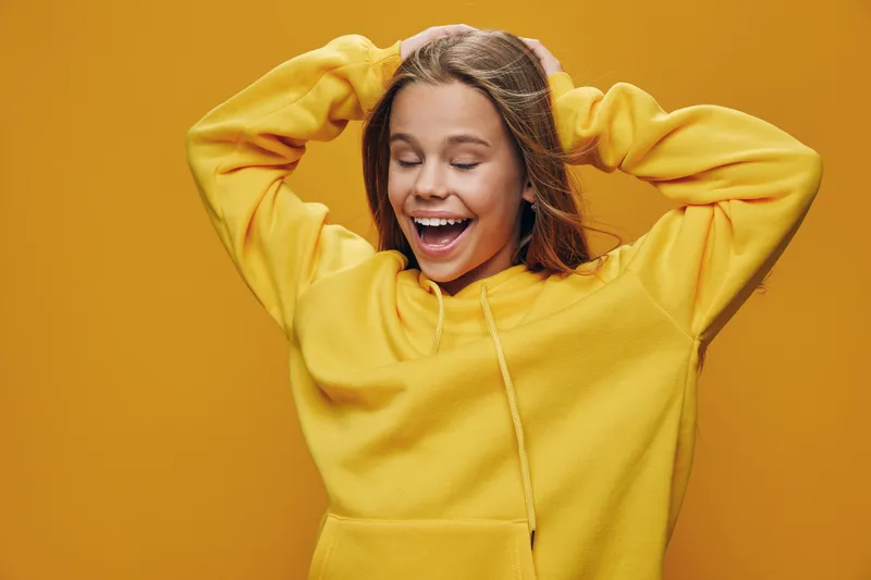 Young woman smiling with closed eyes wearing yellow hoodie on yellow background, hands behind head, happy emotion and casual clothing for lifestyle and youth culture