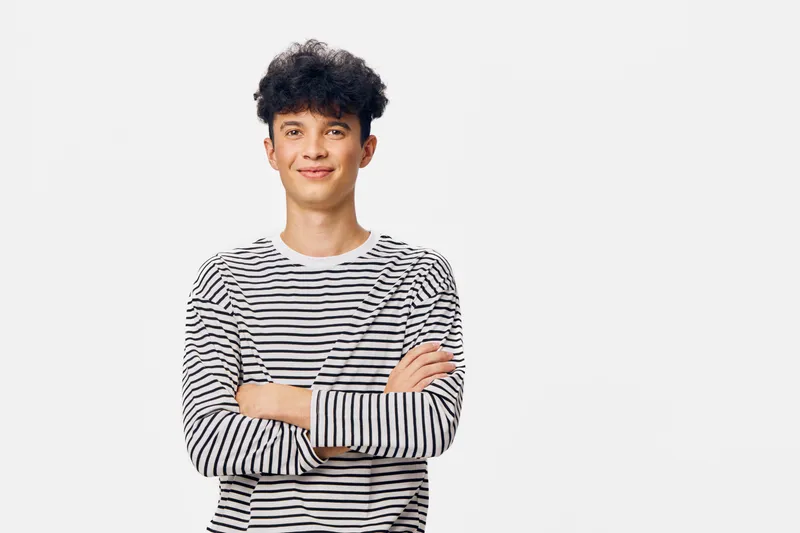 young man wearing striped long sleeve shirt standing with crossed arms, smiling confidently against plain light background, casual fashion style