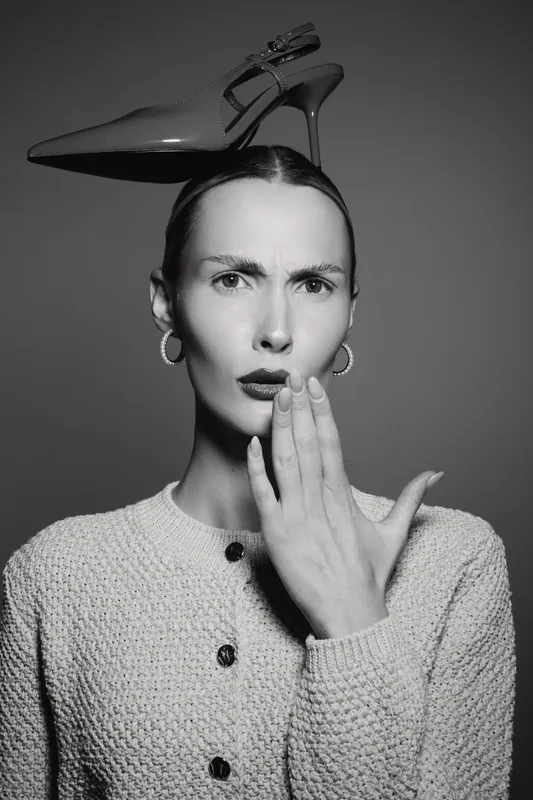 Caucasian woman with pale skin and surprised expression holds hand near mouth, balancing high heel shoe on head.