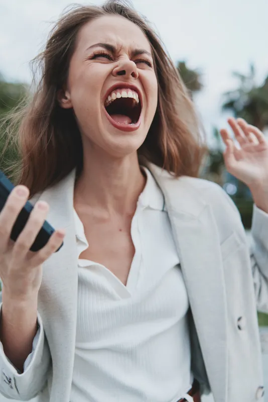 frustrated woman shouting holding smartphone outdoors expressing financial crisis emotions wearing white shirt and light blazer