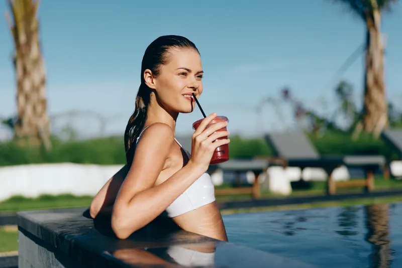 Cheerful woman in a white bikini relaxing at an outdoor pool, sipping a red drink, sunlit moment, calm water, soft breeze, vibrant colors, leisure lifestyle