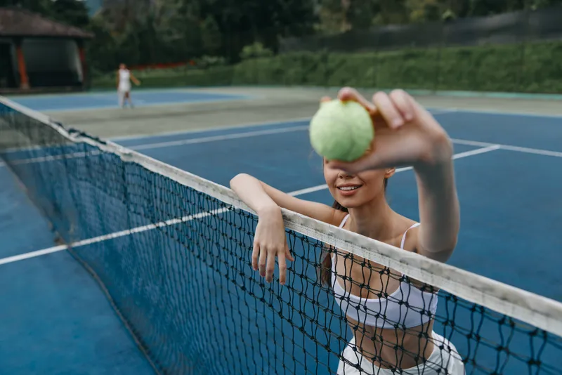 Young woman in sporty outfit smiling and holding a tennis ball on a vibrant blue tennis court, conveying joy and athletic energy during a sunny day