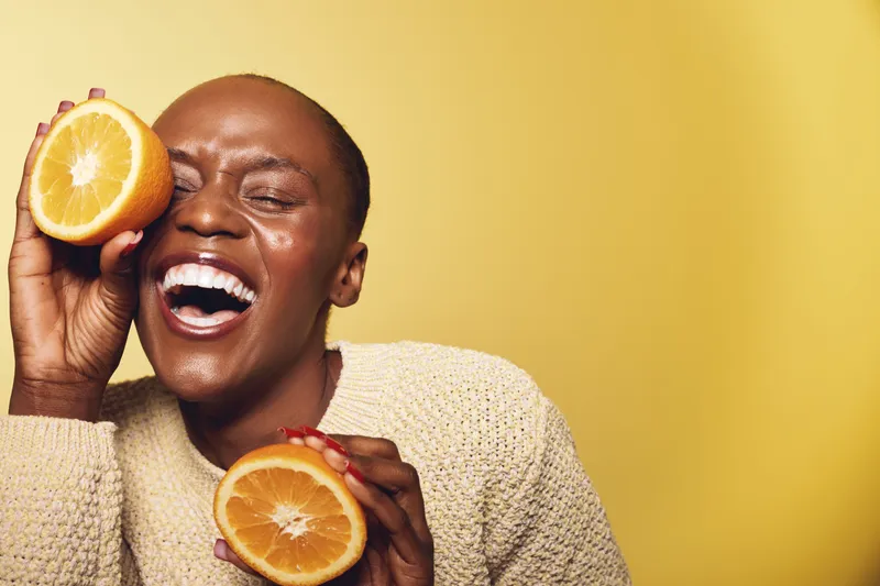 Joyful African woman with dark skin holding two orange halves near her face, laughing with closed eyes. Yellow background and beige sweater create a warm and vibrant mood.