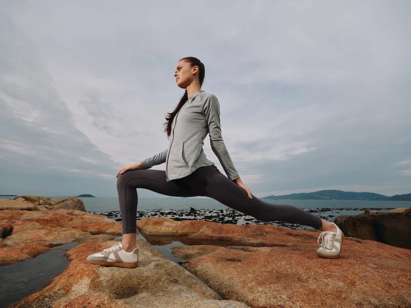 Young athletic woman practicing yoga pose outdoors on rocky beach at sunset, promoting fitness and healthy lifestyle with serene expression