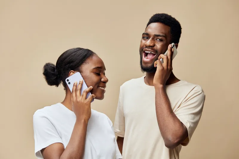 Happy African couple engaging in a playful conversation on mobile phones against a beige background, expressing joy and connection