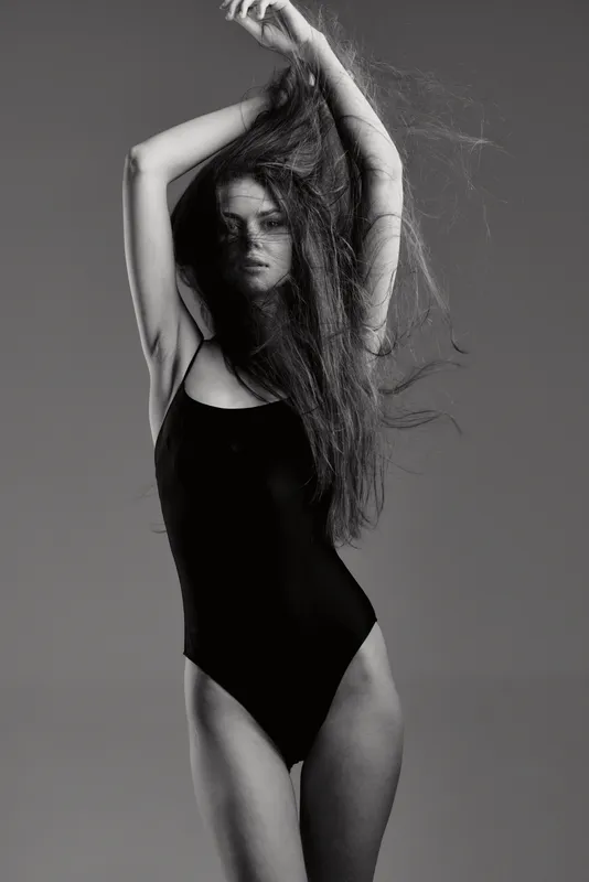 Black and white portrait of woman in black bodysuit with long hair, posing with raised arms, expressive and elegant mood
