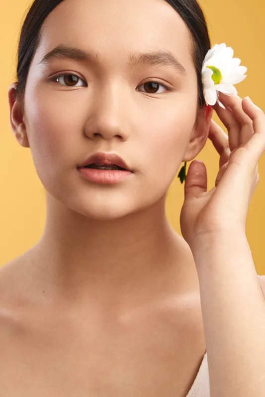 Asian woman presents a serene close up portrait with a flower tucked behind her ear, soft lighting, calm expression, and a warm inviting backdrop that evokes natural beauty and gentle elegance.
