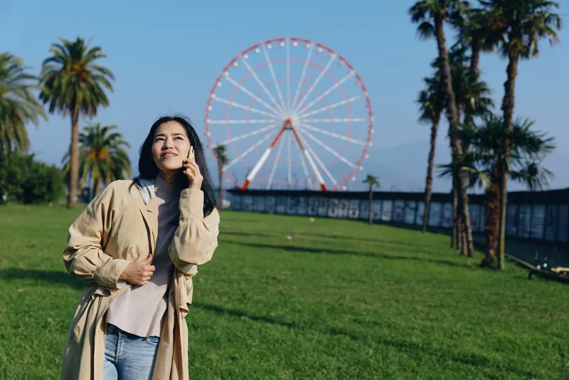 Woman in light beige coat enjoys sunny day in a green park with a distant ferris wheel, casual outdoor moment, travel vibe, leisure scene, blue sky and calm atmosphere