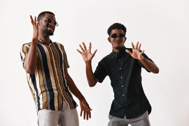 Two young black men with dark skin wearing sunglasses and casual shirts posing and dancing together on white background. Friends having fun and enjoying music in studio.