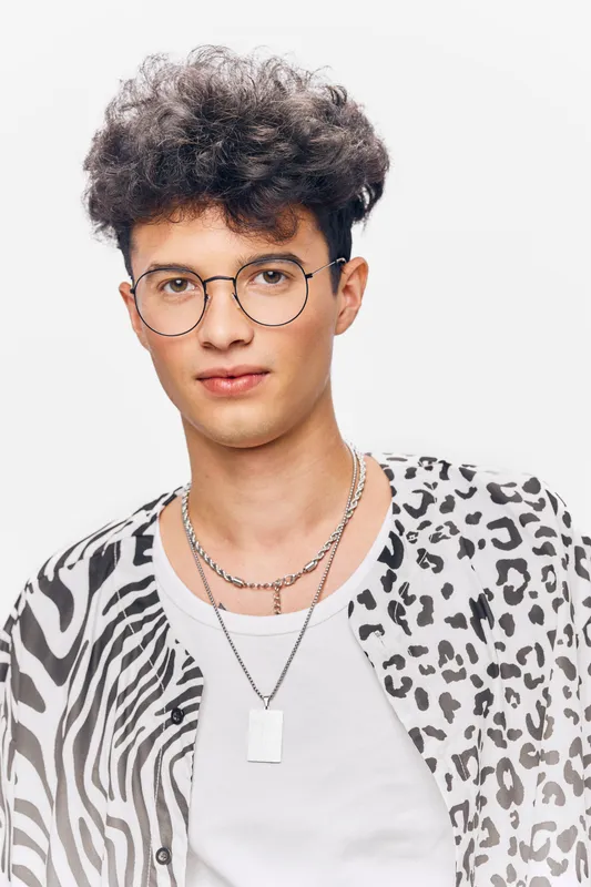 young man with curly hair wearing round glasses and animal print shirt posing confidently on white background, stylish necklace, modern fashion
