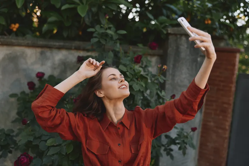 A young woman takes a playful selfie in a garden, wearing a red shirt and smiling as she holds her phone up, enjoying a bright sunny moment in a candid lifestyle shot