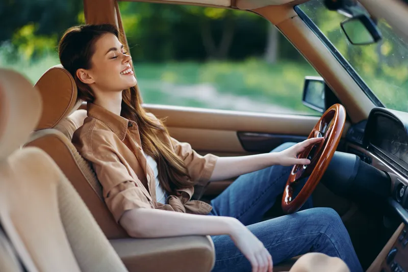 Happy young woman driving vintage car through nature, enjoying summer sunlight, wearing casual attire and displaying a carefree mood on a sunny day.