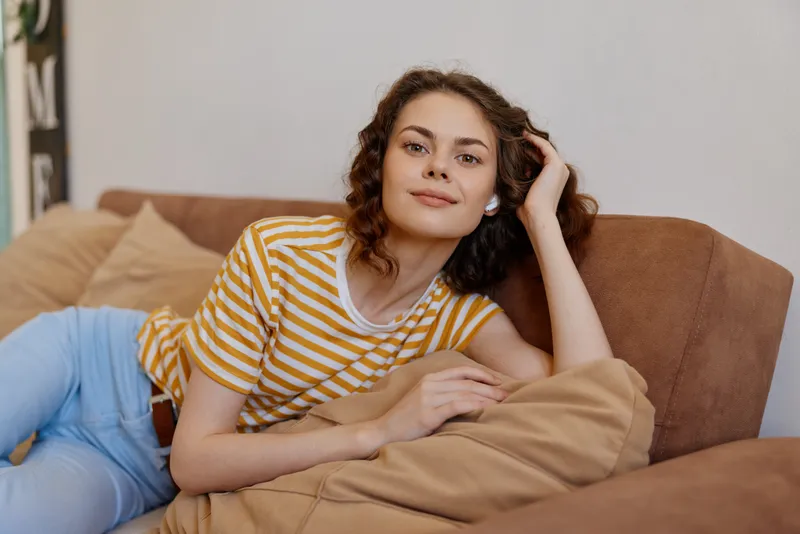 A relaxed young woman sits on a brown couch at home, wearing a yellow striped shirt and light jeans, exuding calm and casual confidence.