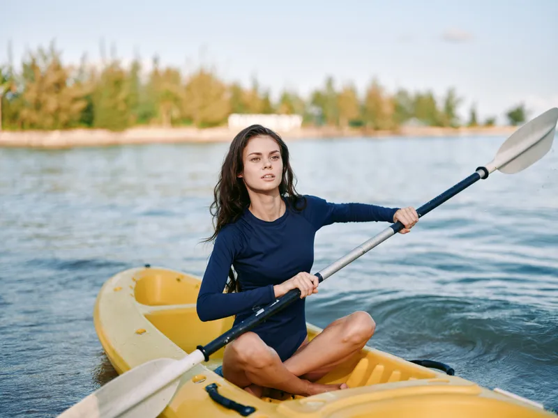 Female kayaker in a navy long sleeve top sits in a yellow kayak on calm water, paddling with a double blade paddle, focused expression, sunlit shoreline in the distance.