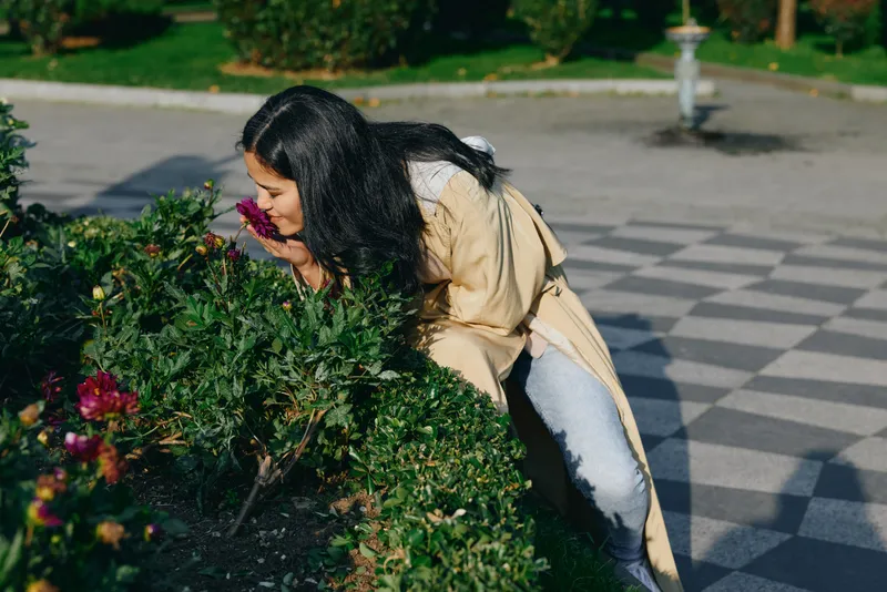 Woman leans toward flowering bushes in a sunlit park, smelling blooms amid nature, outdoor gardening scene, greenery, casual beige coat and jeans, calm, candid moment