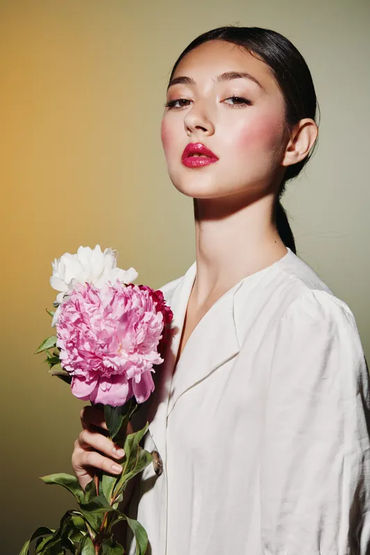 Young woman with natural makeup and glossy red lips holding pink and white peonies on beige and cream background in creative fashion beauty portrait. Soft lighting enhances skin glow.
