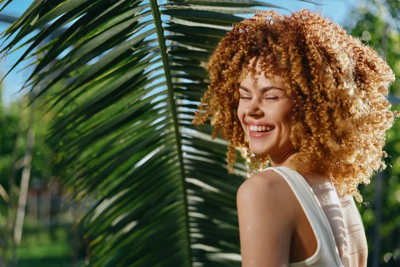 smiling woman with curly hair enjoys sunny day outdoors near large palm leaf, wearing white top, natural daylight, vibrant green background