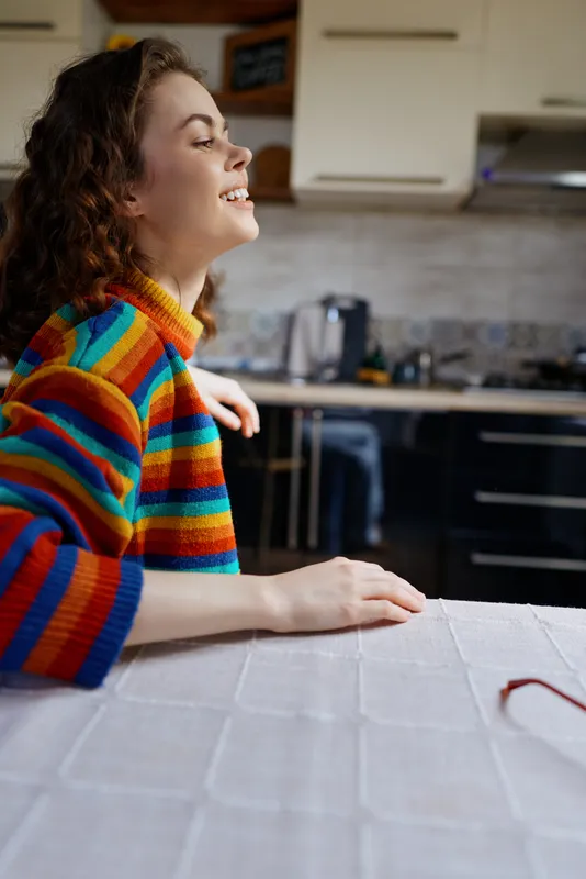 A young woman with curly hair smiles as she rests forearms on a light table in a bright, colorful kitchen, enjoying a calm moment.