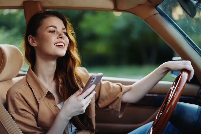 Young woman enjoying a sunny day driving a car, smiling while using her smartphone, wearing casual attire, representing freedom and joy.