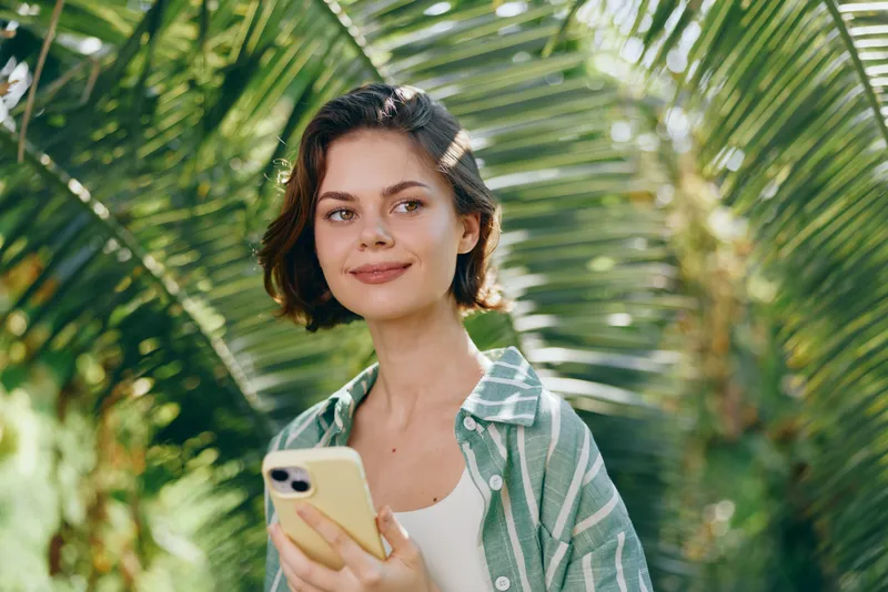 Portrait of a young woman outdoors, smiling softly while holding a smartphone, sunlit tropical backdrop with lush green palm leaves.