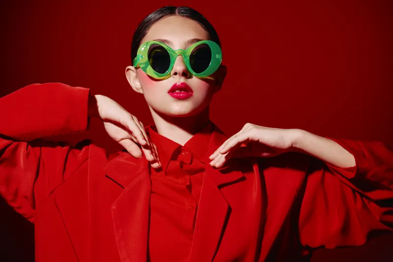 Young woman in oversized green sunglasses wears vivid red coat and lipstick posing stylishly against a red background fashion fashion beauty creative portrait studio modern.