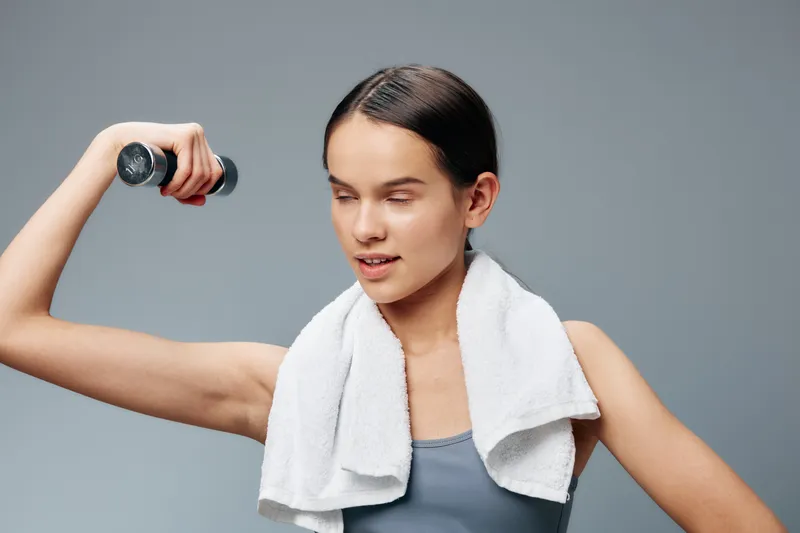 Female fitness enthusiast lifting a dumbbell with a towel around her neck, mid workout in a focused studio setting, showcasing strength, balance, and determination in motion.