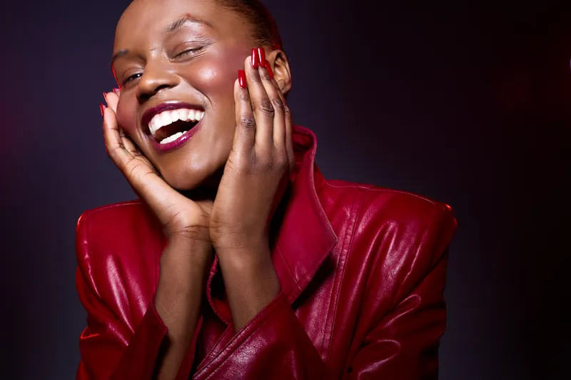 Happy African woman with dark skin wears red leather jacket and smiles joyfully with closed eyes. Studio portrait on dark background. Concept of beauty, fashion, and positive emotions.
