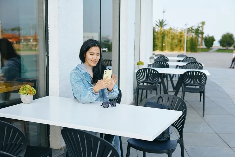 Woman at an outdoor cafe seated at a white table using a smartphone, sunglasses on the table, casual urban setting, daylight, calm moment of daily communication and leisure.