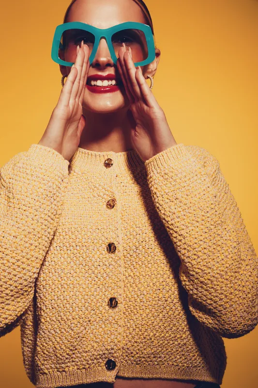 Young woman in knitted sweater and large sunglasses smiles brightly, holding hands near mouth on yellow background. Concept of fashion, style, and joyful expression in studio lighting.