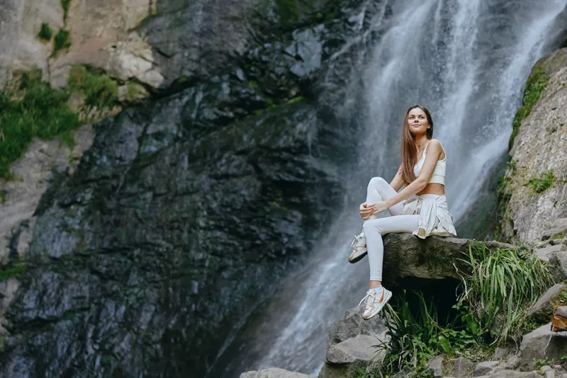 young woman sitting by waterfall in sporty white outfit, enjoying nature, peaceful mood, outdoor, long hair, fresh air, calm environment