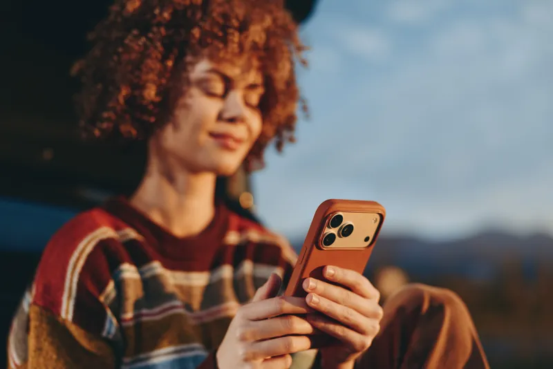Woman lifestyle portrait outdoors, curly haired woman holding smartphone, warm evening light, relaxed expression, using phone for communication, casual clothing, mountain background softly blurred,