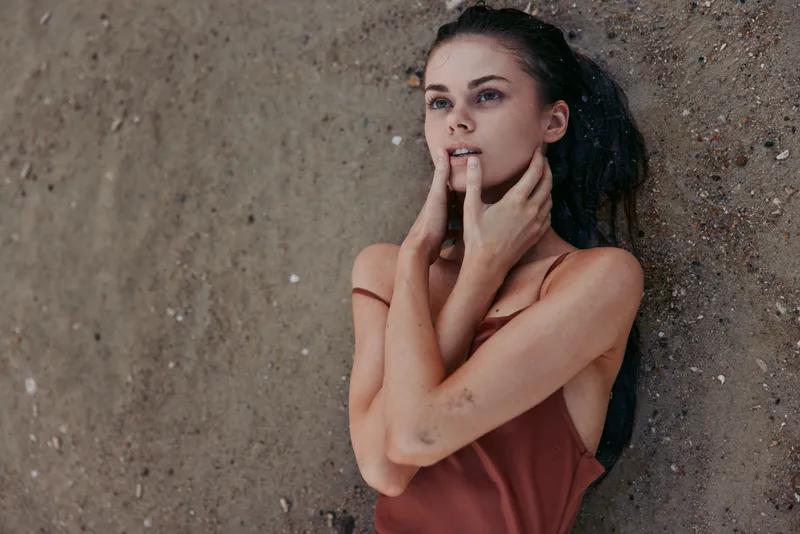 Young woman lies on a rough wall, hands near her mouth, with a pensive gaze and subtle vulnerability. Warm tones, soft light, and an intimate, candid mood define the frame.