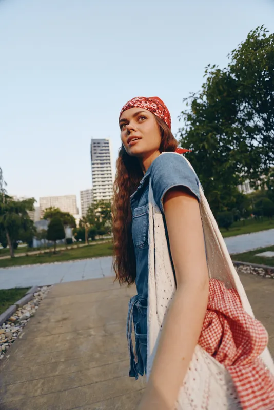 lifestyle woman in modern boho-western denim street style wears red bandana and carries knit bag in natural urban park setting with film color tone
