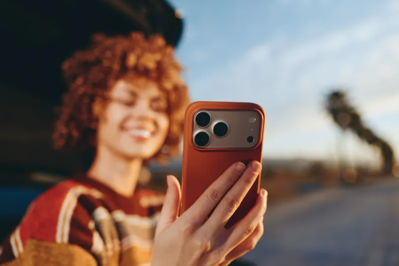 Woman takes a selfie with a smartphone, curly hair framing her face, outdoor lifestyle street tech portrait, smiling while holding the phone, blurred urban background, shallow depth.