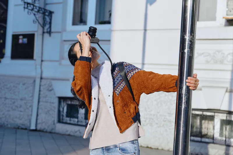 Individual photographer on a sunny urban street raising a camera to shoot, wearing a cozy orange and cream cardigan and denim jeans, with architectural details and soft natural light.