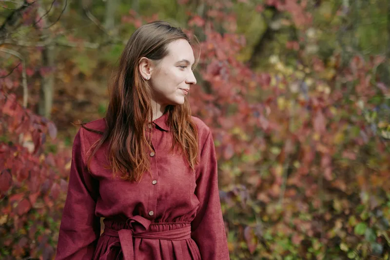 Candid outdoor portrait of a mid-adult woman in a burgundy dress among autumn foliage, capturing a genuine, natural, true-to-life expression and calm presence in warm seasonal light.