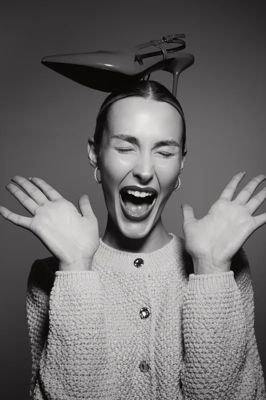 Excited Caucasian woman with fair skin and closed eyes wears high heel shoe on head and laughs loudly.