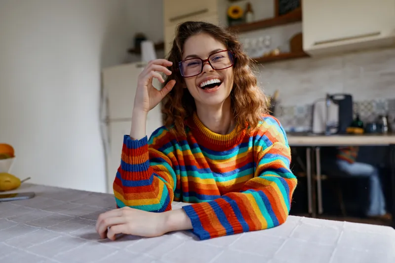 A smiling young woman with curly hair wears a bright striped sweater and sits at a kitchen table, capturing a candid, warm home lifestyle moment in a bright room.