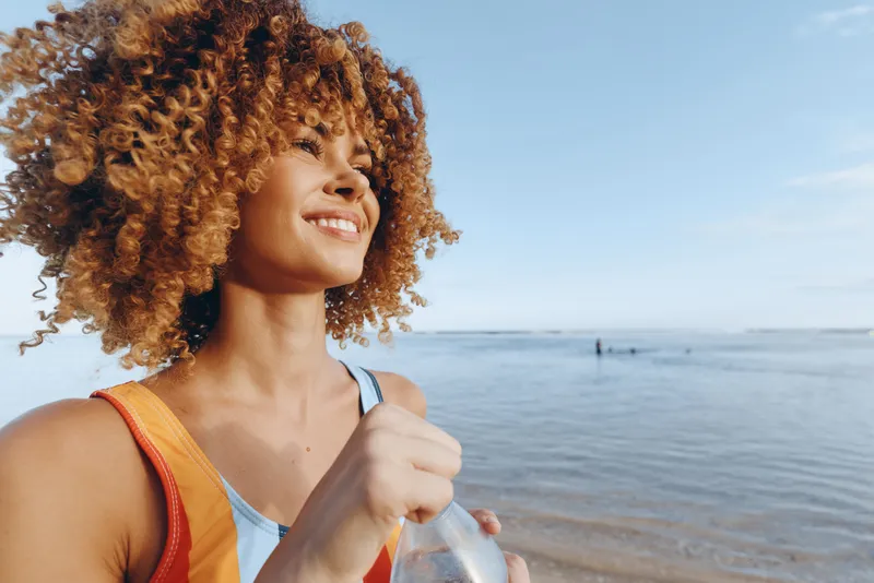 Bright smiling woman on a sunny beach holds a bottle, exuding carefree summer vibes, outdoor portrait with blue sky, calm sea, golden sand, and vibrant hairstyle.