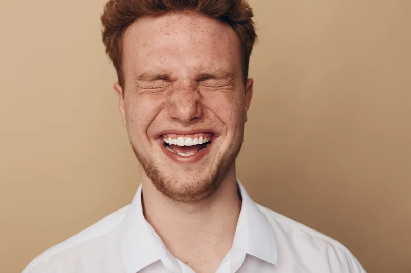Happy young man with red hair and freckles laughs with eyes closed wearing white shirt on beige background. Concept of joy, positive emotions, and natural expression.