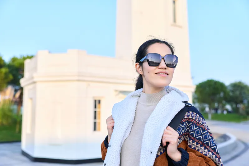 Model wearing oversized sunglasses and layered chic outerwear stands outdoors beside a light monument, showcasing fashion, confidence, and sunny day style with cinematic lighting and modern