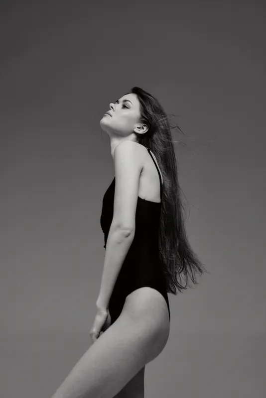 Black and white portrait of a young woman in a black swimsuit standing gracefully with closed eyes and long hair flowing calmly.