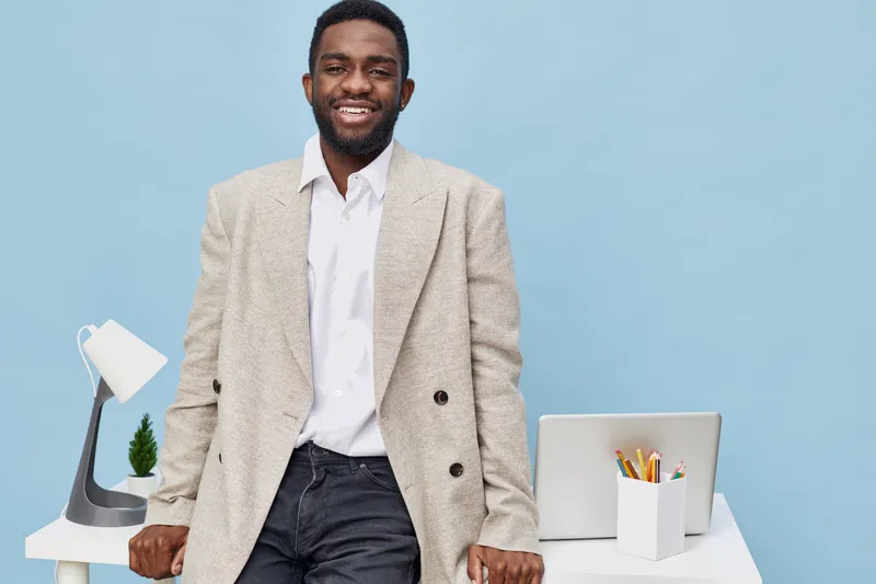 Confident smiling man standing in modern office interior with white desk and accessories, dressed in smart casual suit, posing against pastel blue background, workplace, personality, professionalism c