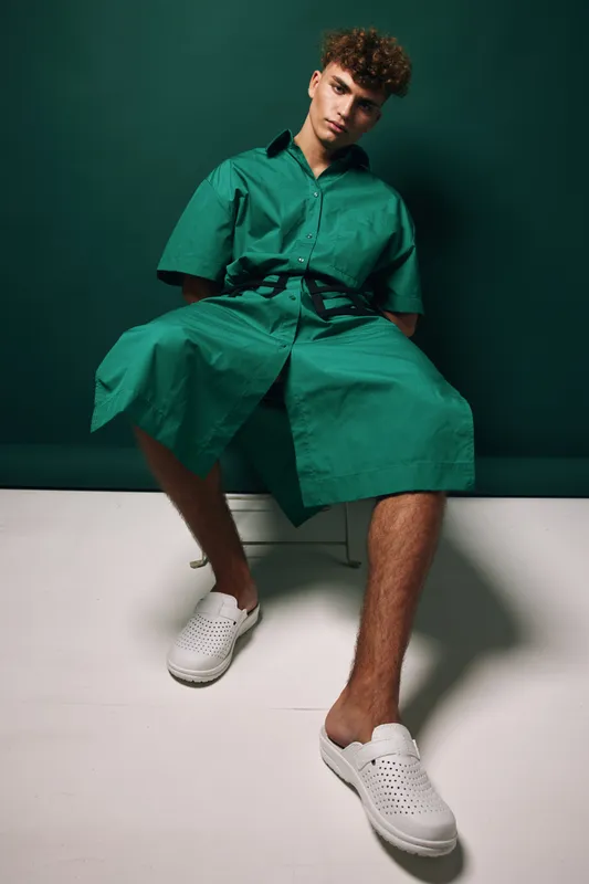Young man sitting and wearing oversized green shirt dress, casual minimalist fashionable look with white slip on shoes and curly hair in modern studio against dark green wall background.