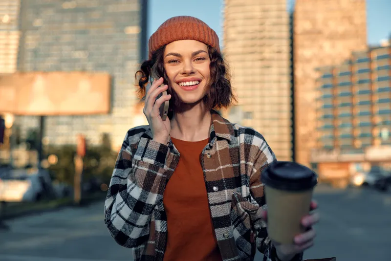 Woman smile coffee phone urban city beanie sunlight candid lifestyle portrait of a young female in plaid jacket, golden hour glow, authenticity, mindful living and emotional storytelling.