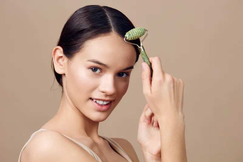 Young woman using a jade roller on her face for skincare, showcasing a healthy glow and natural beauty in a neutral backdrop, perfect for wellness themes