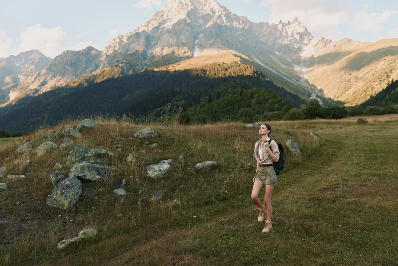 Young woman hiking in mountains wearing backpack and shorts enjoys nature landscape with rocky peaks and green forest under clear sky during daytime.